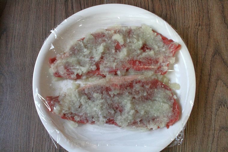 白い皿にすりおろし玉ねぎを塗った牛ステーキ肉が置かれラップをした様子