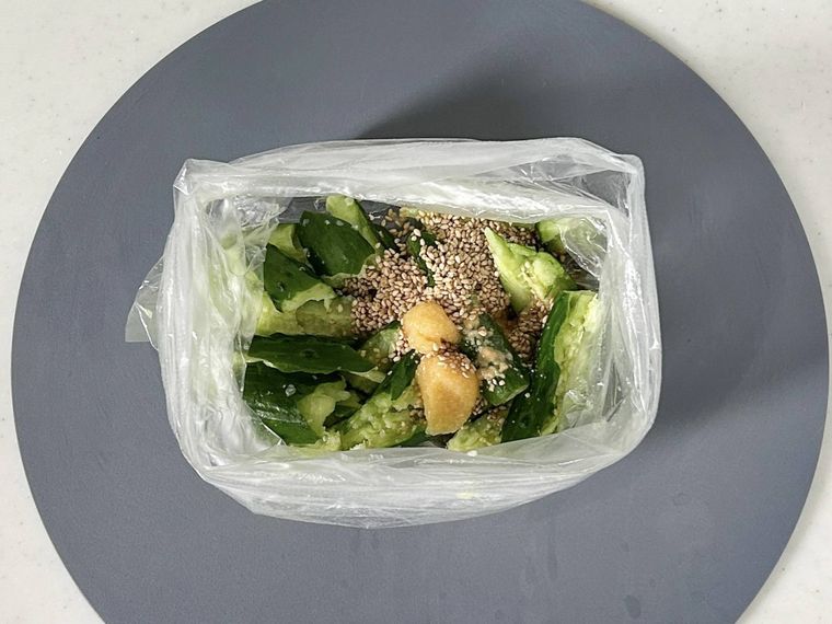 グレーのまな板の上に置かれているポリ袋に入ったきゅうり、鶏ガラスープの素、レモン果汁、ごま油、白いりごま、おろしにんにく