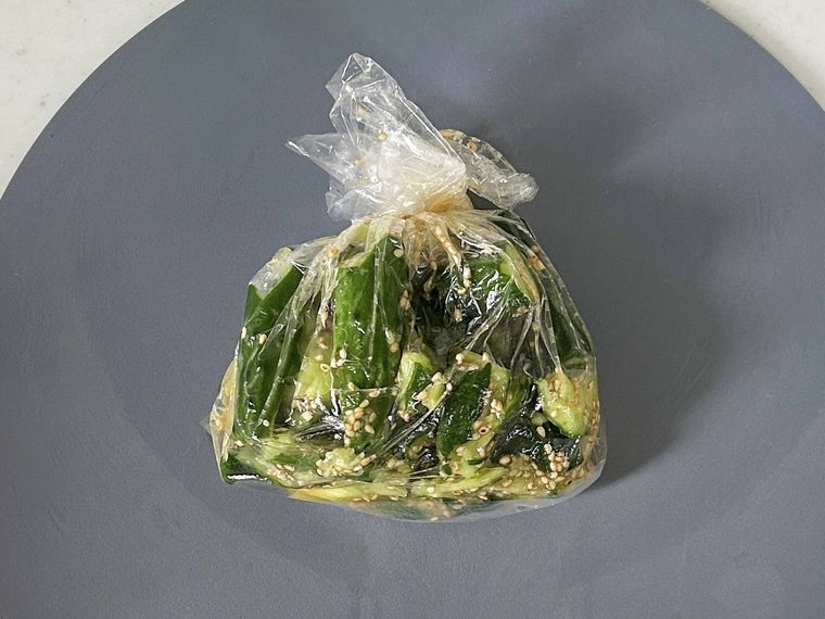 グレーのまな板の上に置かれているポリ袋に入ったきゅうり、鶏ガラスープの素、レモン果汁、ごま油、白いりごま、おろしにんにく