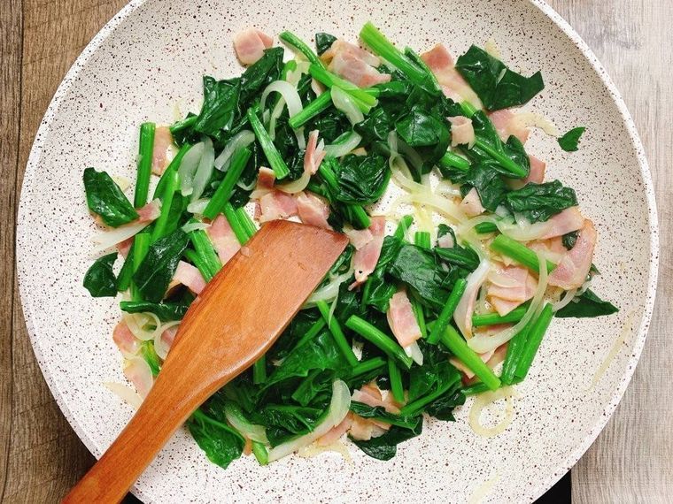 白のフライパンで、ほうれん草、ベーコン、玉ねぎを炒めている様子