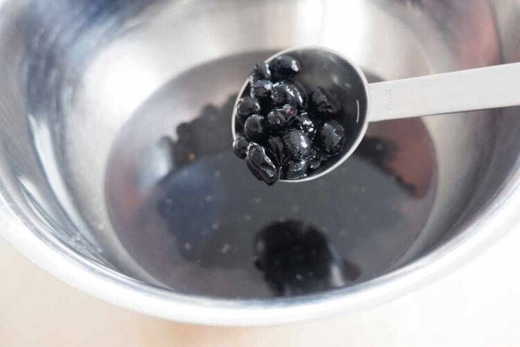 銀のボウルに入った水に計量スプーンで黒豆を加える