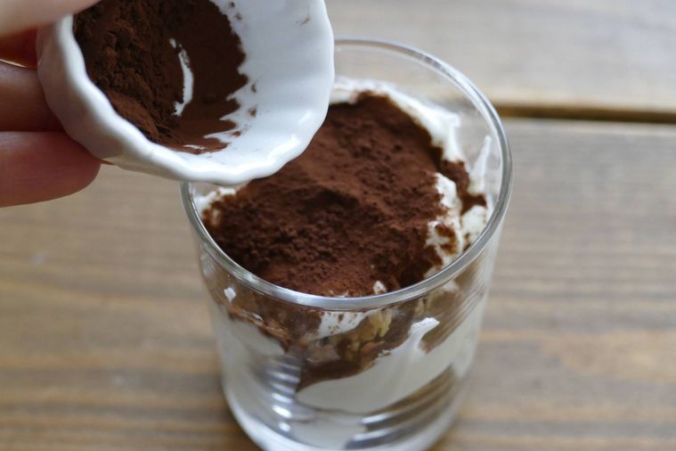 グラノーラとギリシャヨーグルトを重ねたグラスに純ココアパウダーをかける様子