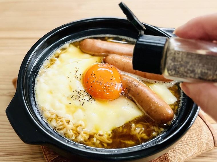 ウインナー、チーズ、卵黄がのったチキンラーメンに粗挽き黒こしょうを振っている様子