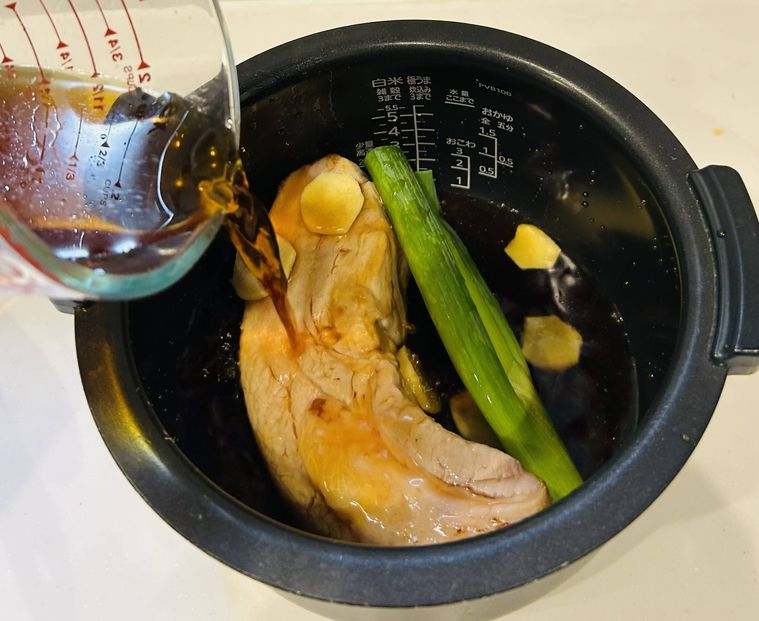 炊飯器の内釜に豚バラ肉、長ねぎ、しょうがを入れ、タレを注いでいる様子